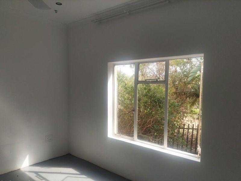 A bright, empty bedroom featuring white walls, a ceiling fan, and a large window overlooking the garden.