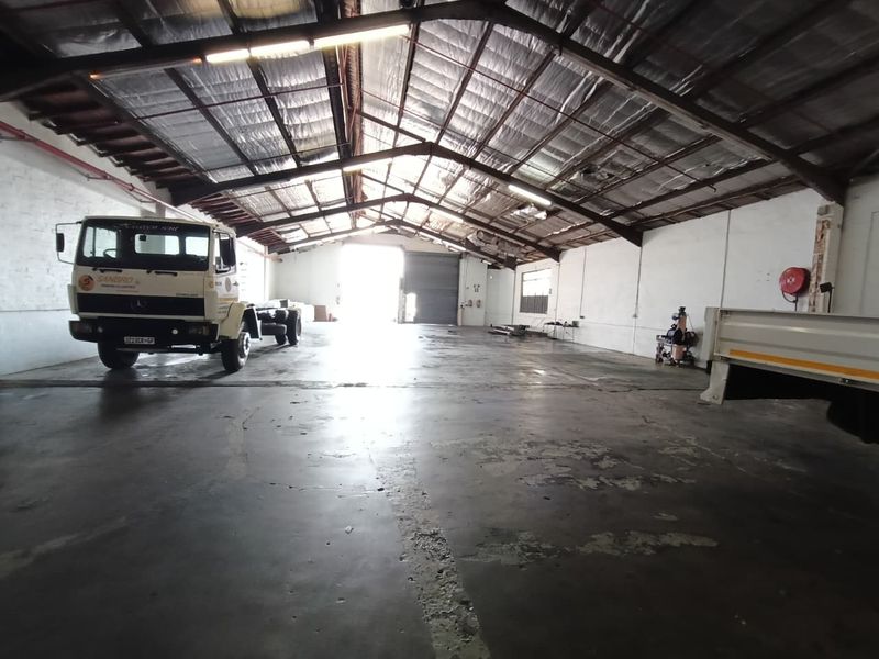 Spacious industrial warehouse or garage featuring high ceilings with exposed steel trusses, concrete flooring, and a large roller shutter door for vehicle access.