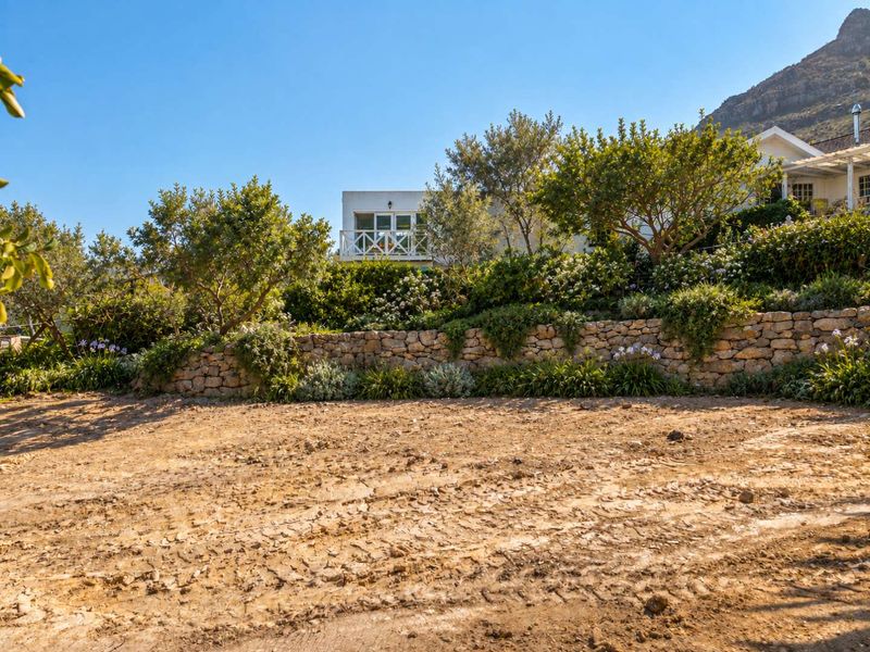 A view of a vacant residential plot featuring a stone retaining wall and established garden landscaping with mature trees.