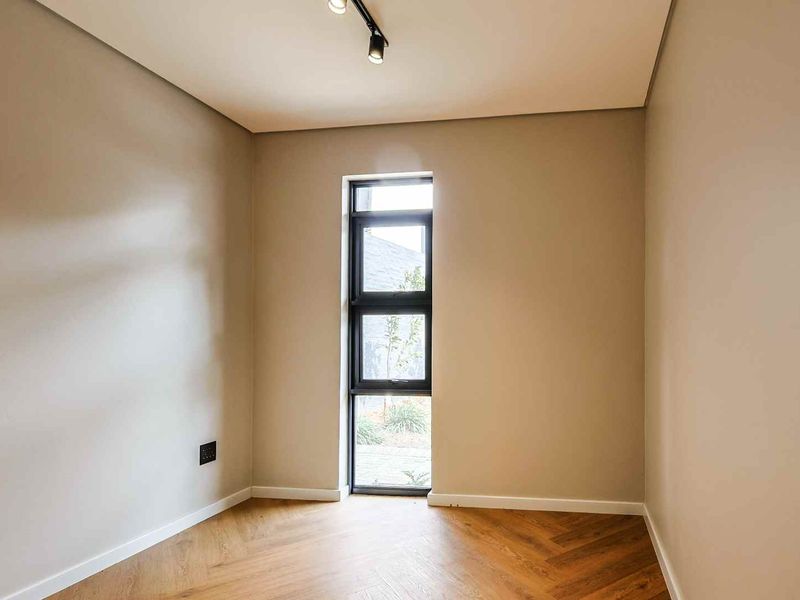 A bright, empty bedroom featuring modern herringbone wood flooring, neutral walls, and a tall black-framed window providing natural light.