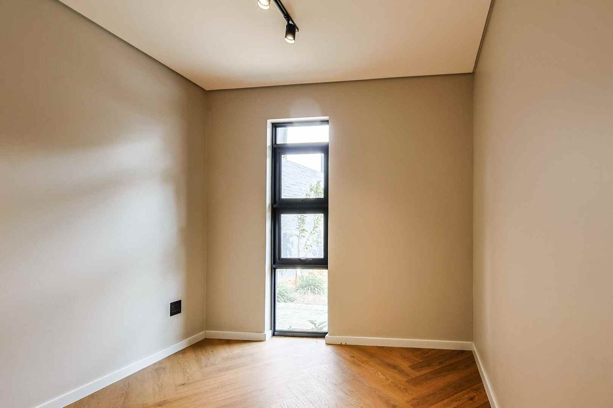 A bright, empty bedroom featuring modern herringbone wood flooring, neutral walls, and a tall black-framed window providing natural light.