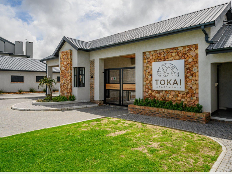 The entrance to Tokai Apartments featuring modern architecture with stone cladding, a metal roof, and a secure gated access point.