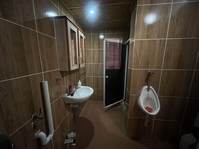 A functional bathroom featuring full-height wall tiling, a wall-mounted washbasin, and a urinal, with exposed plumbing and dated finishes throughout.