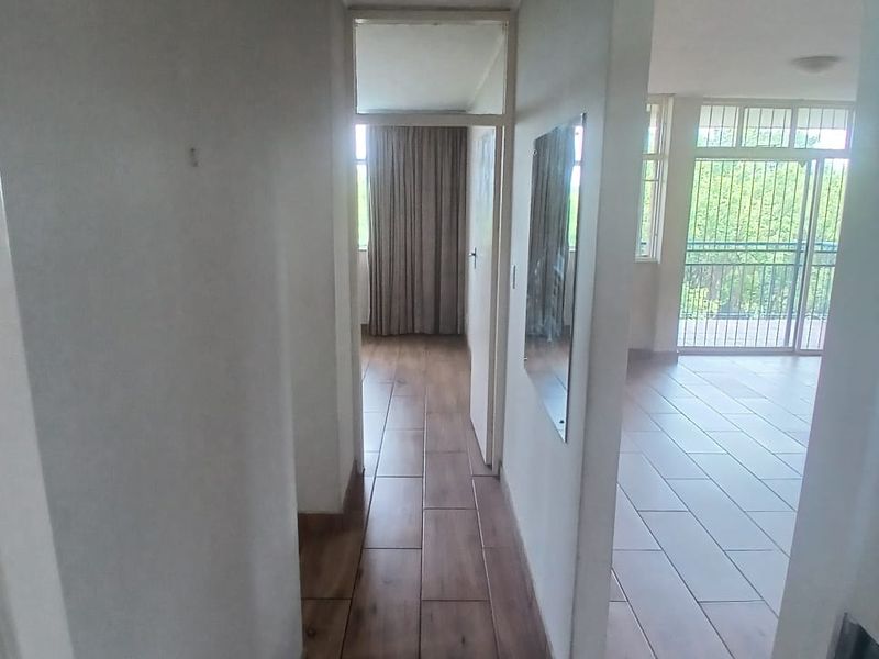 A bright, empty hallway featuring wood-look tiled flooring, leading towards a living area with access to a balcony through a security gate.