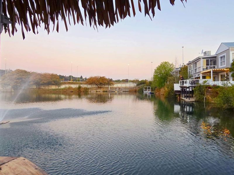 A scenic view overlooking a tranquil lake with a water fountain, featuring a thatched-roof structure in the foreground and residential properties along the bank.