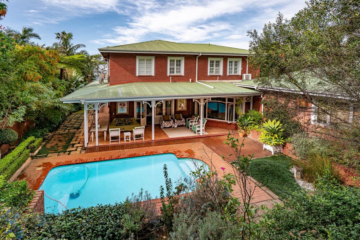 Rear view of a two-story brick house featuring a covered patio, timber decking, and a swimming pool set within a landscaped garden.