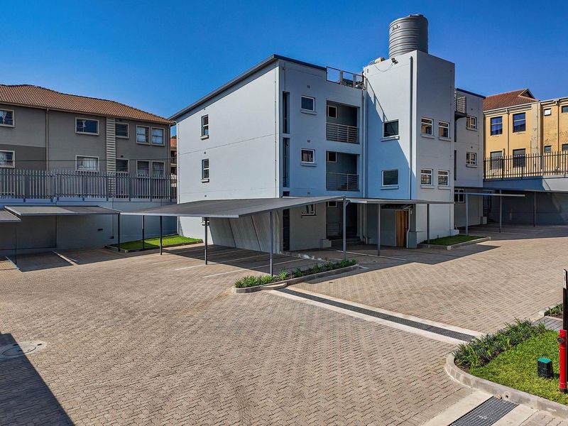 Exterior view of the apartment complex featuring a paved parking area with covered carports, perimeter fencing, and a rooftop water storage tank.