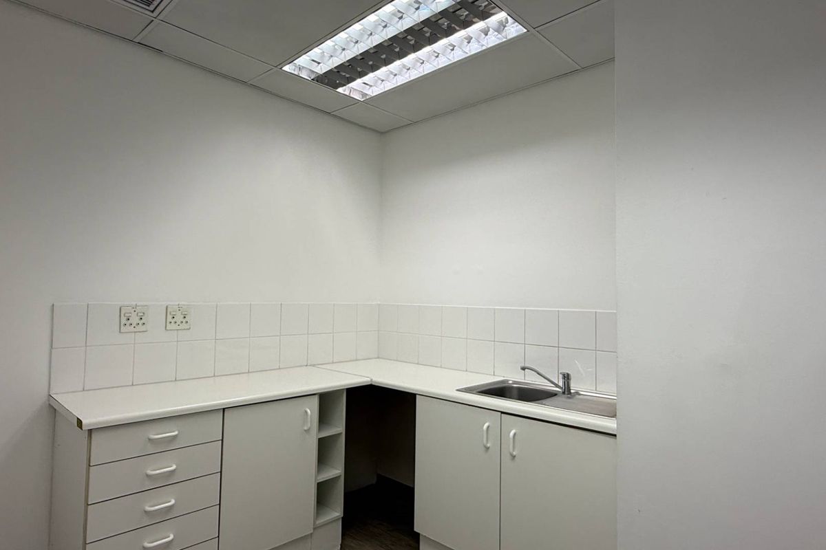 A clean, functional office kitchenette featuring white cabinetry, a stainless steel sink, tiled backsplash, and recessed ceiling lighting.