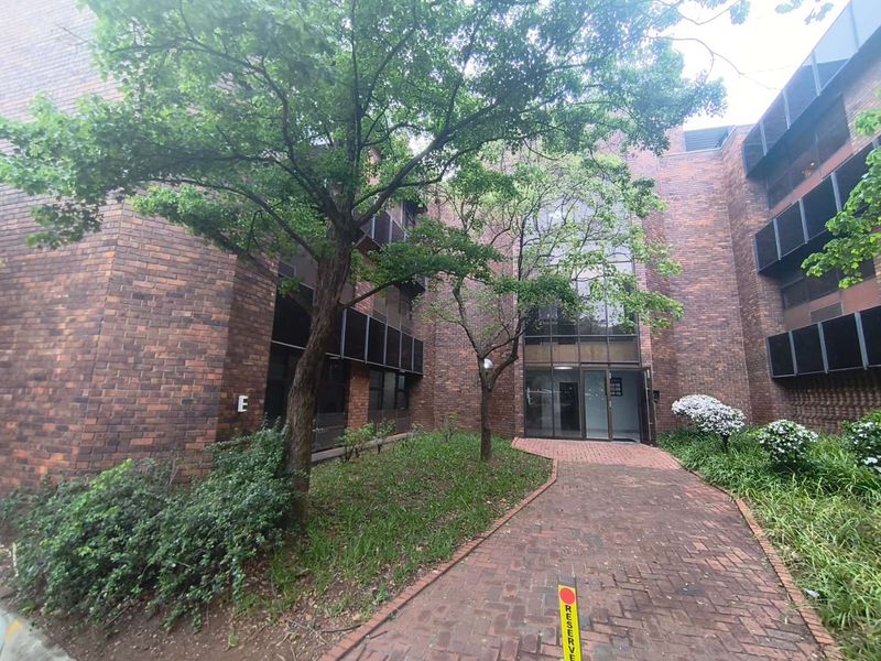 Front exterior view of a multi-story office building featuring a brick facade, large glass entrance, and a paved walkway through a landscaped garden area.