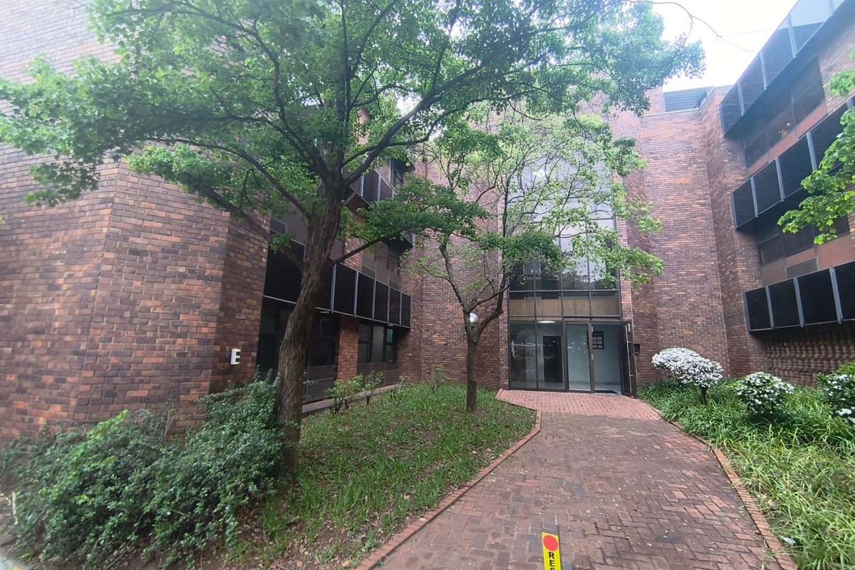 Front exterior view of a multi-story office building featuring a brick facade, large glass entrance, and a paved walkway through a landscaped garden area.