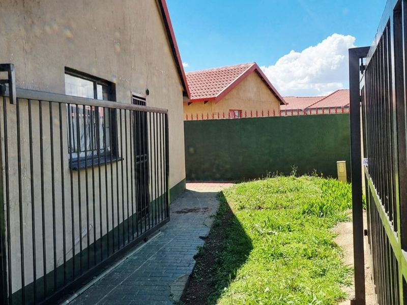 A side view of the property featuring a paved walkway, metal security fencing, and a small grassed area enclosed by a perimeter wall.