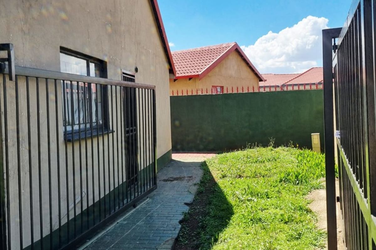 A side view of the property featuring a paved walkway, metal security fencing, and a small grassed area enclosed by a perimeter wall.