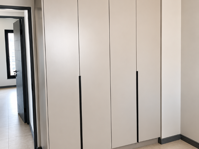 A modern bedroom featuring sleek, floor-to-ceiling built-in wardrobes with minimalist handles, neutral tiled flooring, and recessed ceiling lighting.