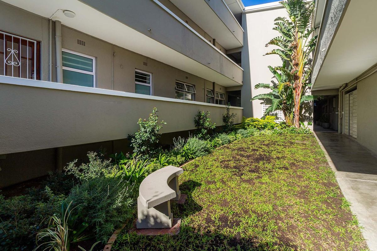 A well-maintained communal garden area within an apartment complex, featuring landscaped greenery, a concrete pathway, and secure window access.