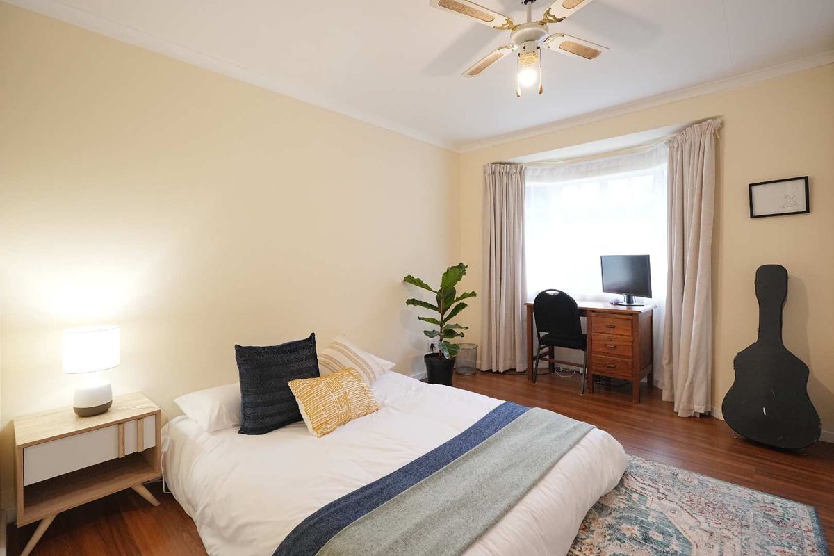 A well-lit bedroom featuring warm-toned wood flooring, a ceiling fan, and a bay window with curtains.