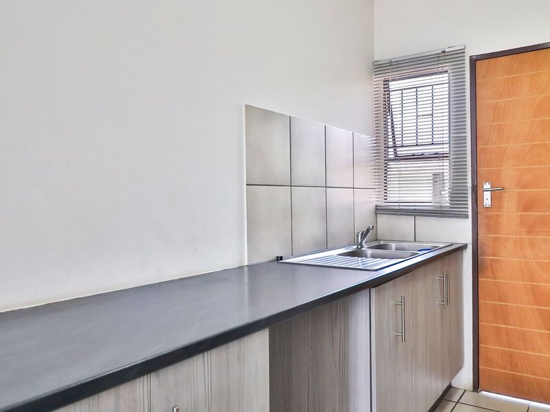 A functional kitchen area featuring a long laminate countertop, stainless steel sink, tiled splashback, and built-in cabinetry with a wooden door.