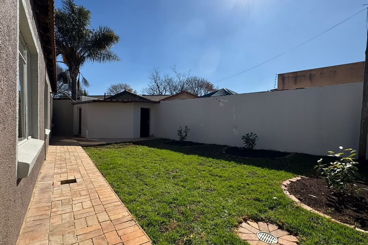 A rear exterior view featuring a paved walkway, a small lawn area, and a boundary wall with an adjacent outbuilding.
