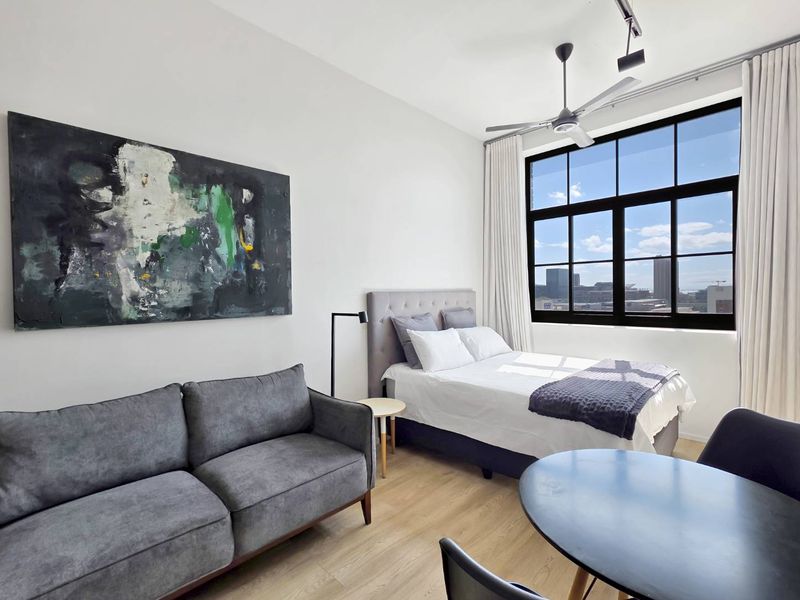 A bright, modern bedroom featuring high ceilings, large industrial-style windows with city views, and durable laminate flooring.