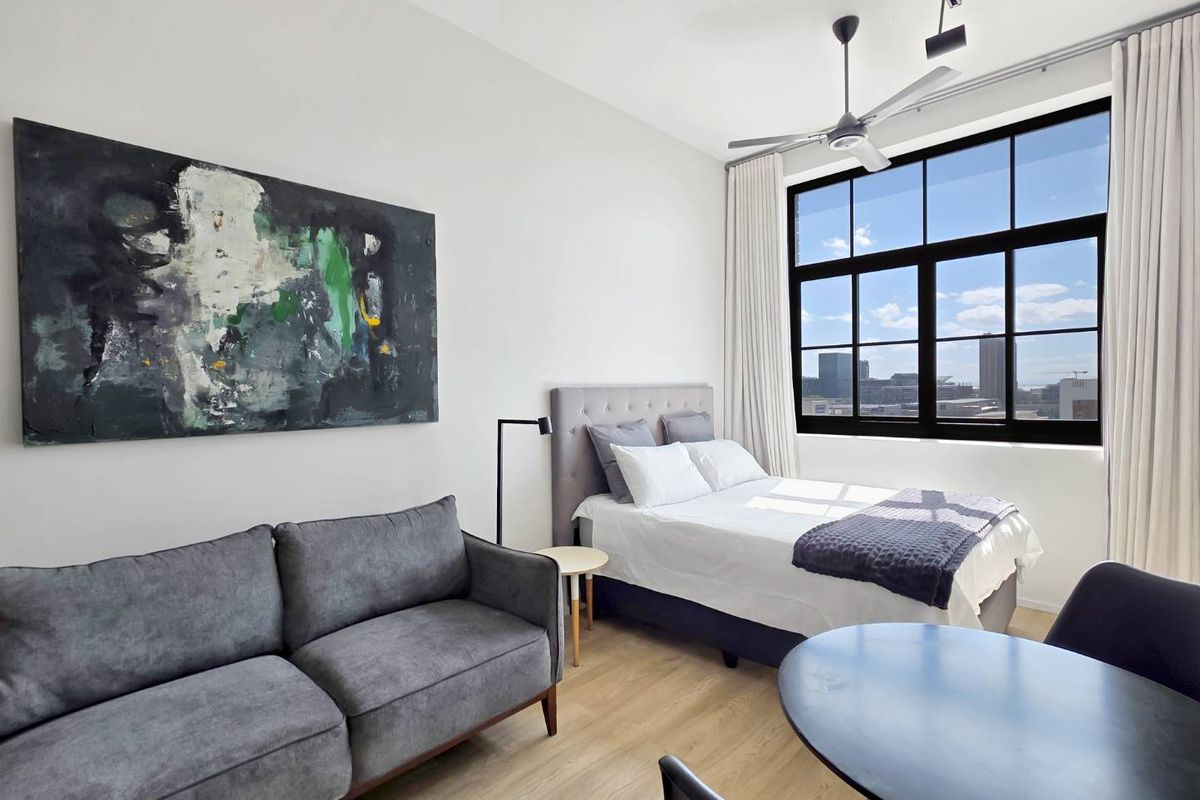 A bright, modern bedroom featuring high ceilings, large industrial-style windows with city views, and durable laminate flooring.