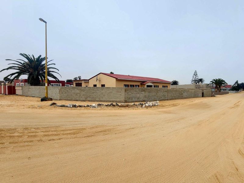 Street view of a residential property featuring a perimeter wall and metal roof, situated on an unpaved sandy road.