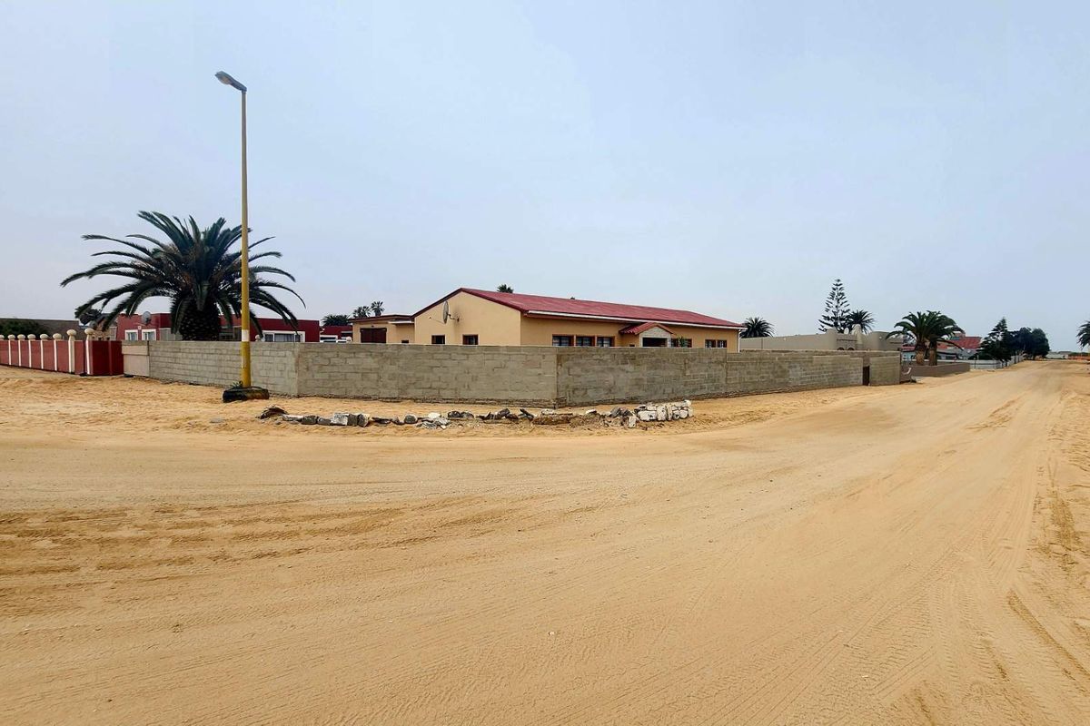 Street view of a residential property featuring a perimeter wall and metal roof, situated on an unpaved sandy road.