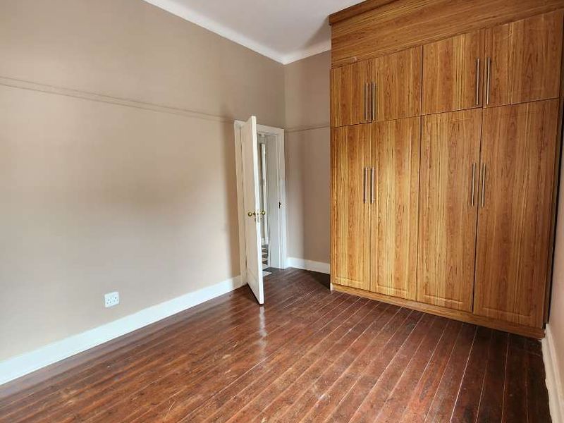 A bright, empty bedroom featuring polished hardwood flooring, a large built-in wooden wardrobe, and a classic ceiling light fixture.