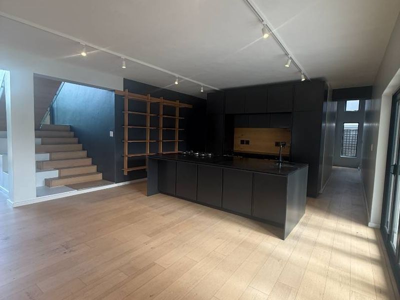 Modern open-plan kitchen featuring dark cabinetry, a central island, and light hardwood flooring, adjacent to a staircase in a bright, contemporary space.