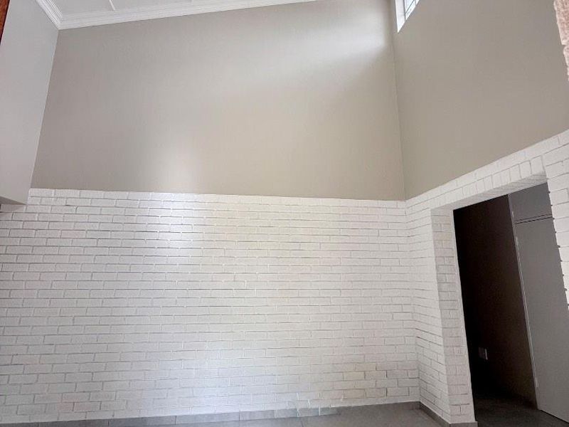 A bright, spacious living area featuring high vaulted ceilings, a modern pendant light, tiled floors, and a distinctive white-painted exposed brick feature wall.