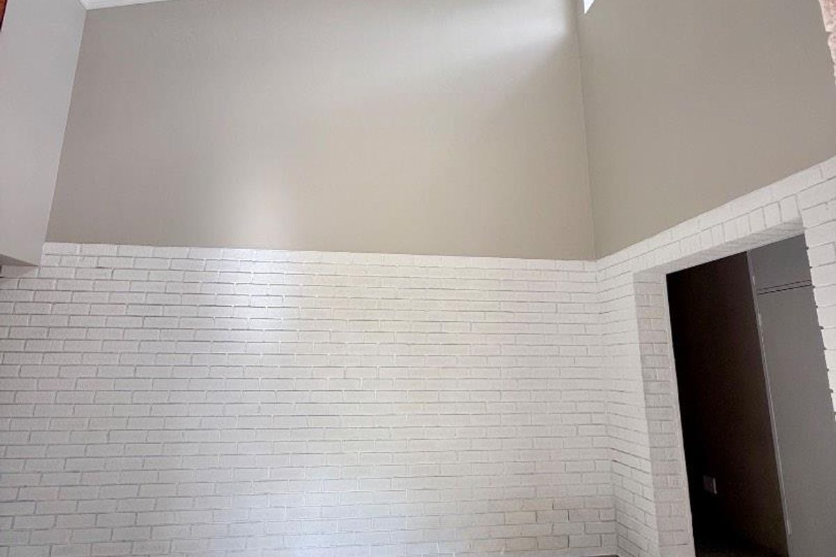 A bright, spacious living area featuring high vaulted ceilings, a modern pendant light, tiled floors, and a distinctive white-painted exposed brick feature wall.