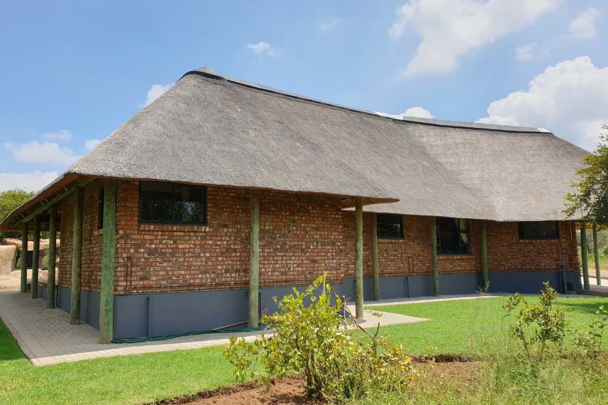 A well-maintained brick building featuring a traditional thatched roof, wooden support pillars, and a paved walkway set within a landscaped garden area.