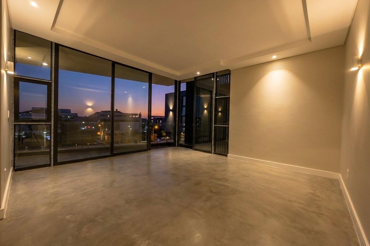 A modern, empty living room featuring polished concrete floors, recessed ceiling lighting, and expansive floor-to-ceiling glass sliding doors with sunset views.