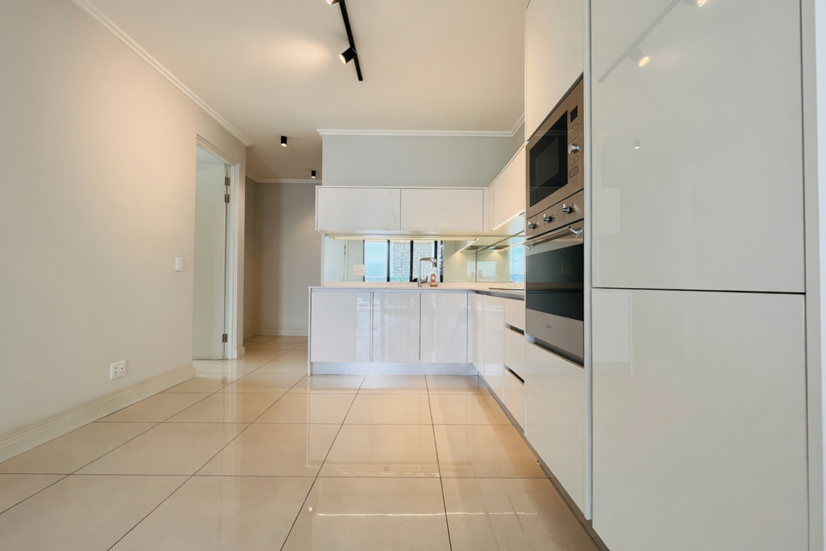 Modern kitchen featuring high-gloss white cabinetry, integrated oven and microwave, mirrored splashback, and sleek tiled flooring in an open-plan layout.