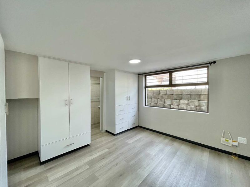 A bright, empty bedroom featuring modern laminate flooring, ample built-in cupboard storage, and a large window with a view of a retaining wall.