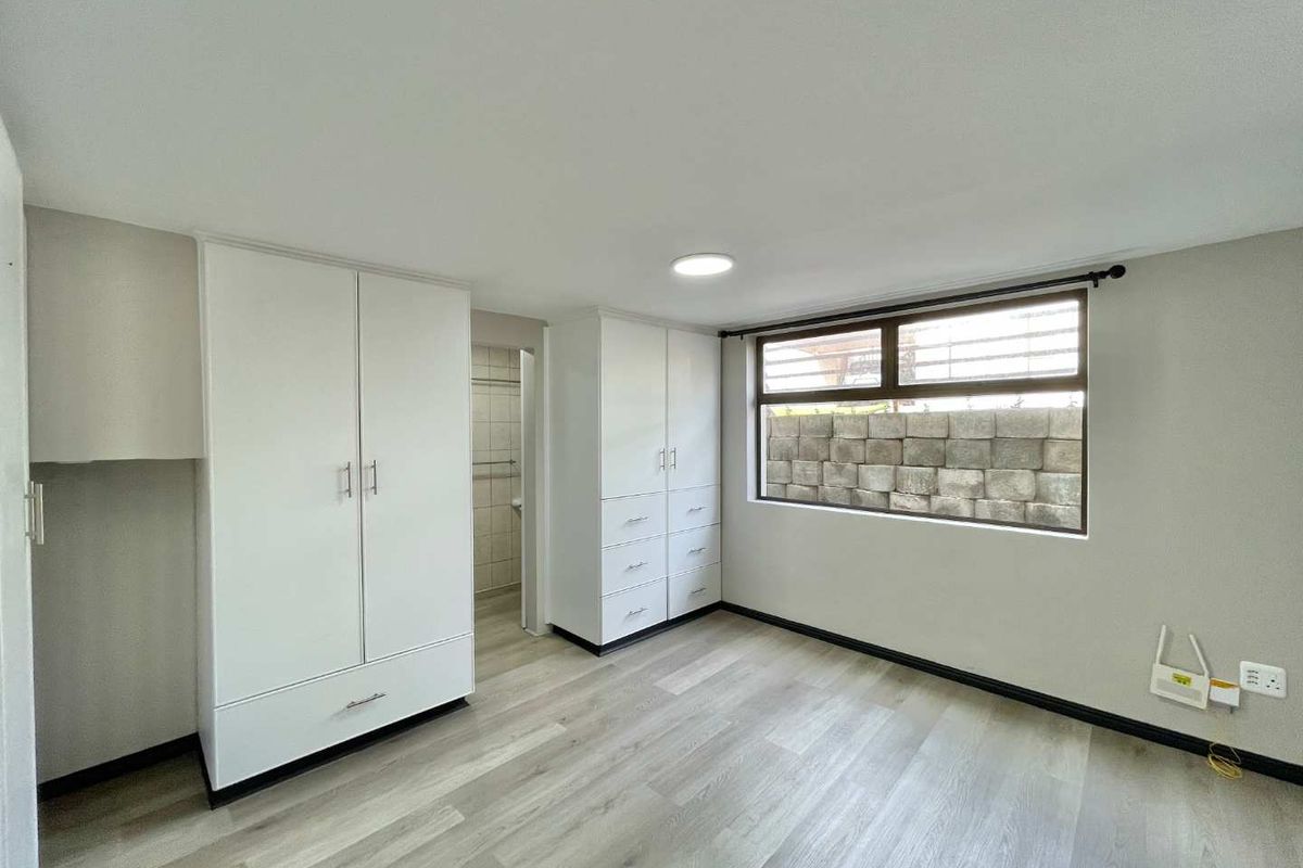 A bright, empty bedroom featuring modern laminate flooring, ample built-in cupboard storage, and a large window with a view of a retaining wall.