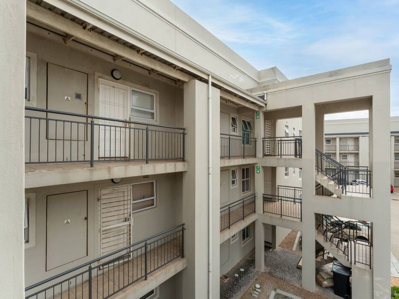 Exterior view of an apartment complex featuring concrete walkways, metal balustrades, and a central stairwell leading to a paved ground-level courtyard.