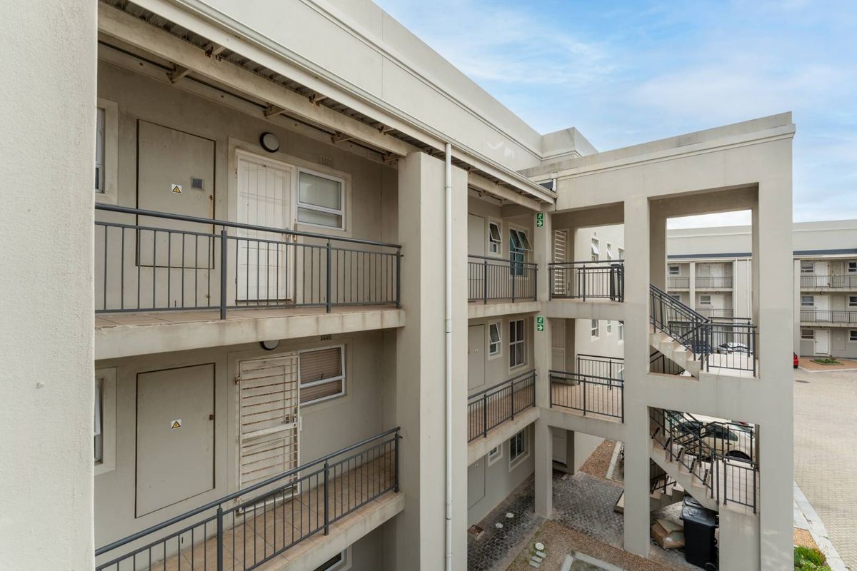 Exterior view of an apartment complex featuring concrete walkways, metal balustrades, and a central stairwell leading to a paved ground-level courtyard.