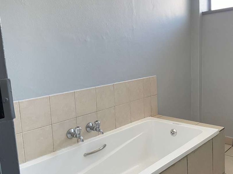 A functional bathroom featuring a built-in bathtub with tiled surround and splashback, ceramic floor tiles, and a window with security bars.