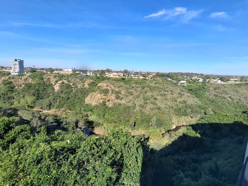 An elevated scenic view overlooking a lush green valley with natural vegetation and distant residential buildings under a clear blue sky.