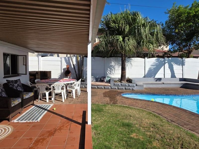 A covered patio area overlooking a swimming pool, featuring brick paving, a built-in braai, and a landscaped garden with perimeter walling.