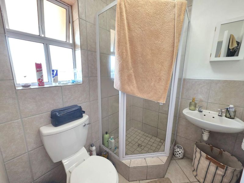 A functional bathroom featuring a corner shower enclosure, pedestal washbasin, toilet, and neutral ceramic tiling throughout.