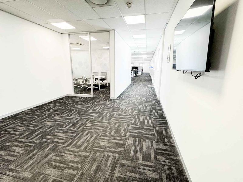 A modern office hallway featuring carpeted flooring, suspended ceiling with recessed lighting, and a glass partition wall leading to an adjacent workspace.