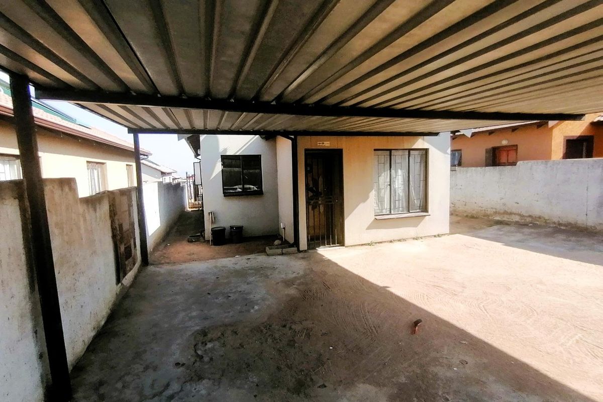 Rear exterior area featuring a corrugated metal carport, concrete flooring, and perimeter walling with security gate and burglar bars on windows.