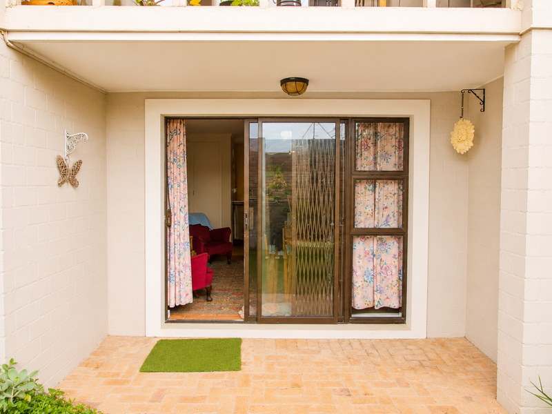A covered patio area featuring brick paving and a sliding glass door with a security gate providing access to the interior living space.