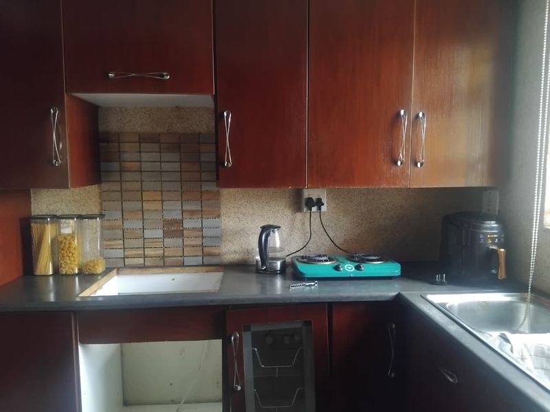 A compact kitchen featuring dark wood-look cabinetry, a mosaic tile backsplash, and a stainless steel sink, currently showing signs of wear and missing cabinetry components.