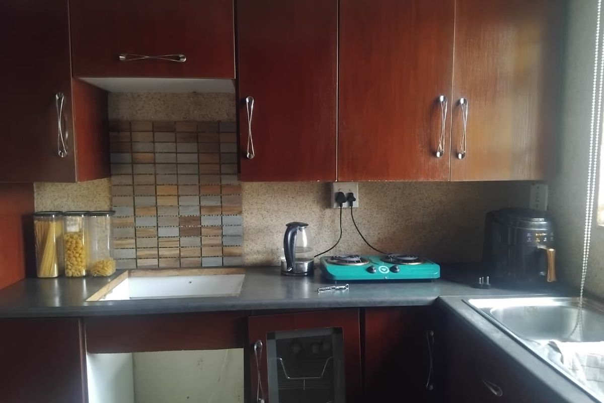 A compact kitchen featuring dark wood-look cabinetry, a mosaic tile backsplash, and a stainless steel sink, currently showing signs of wear and missing cabinetry components.