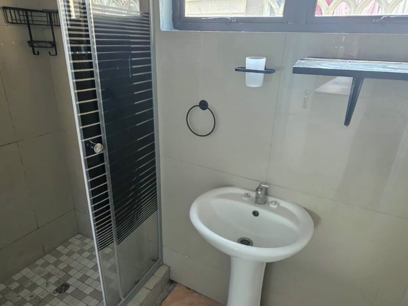 A functional bathroom featuring a pedestal basin, a glass-enclosed shower, and neutral wall tiling. The space is clean but requires minor finishing touches.
