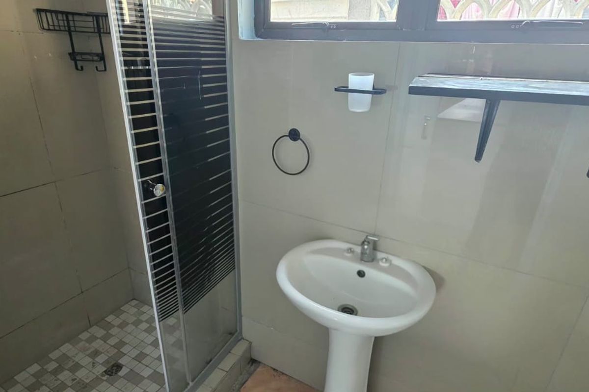 A functional bathroom featuring a pedestal basin, a glass-enclosed shower, and neutral wall tiling. The space is clean but requires minor finishing touches.