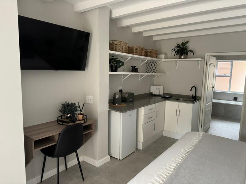 A compact kitchenette featuring white cabinetry, stone countertops, open shelving, and exposed ceiling beams, seamlessly integrated into a modern living space.