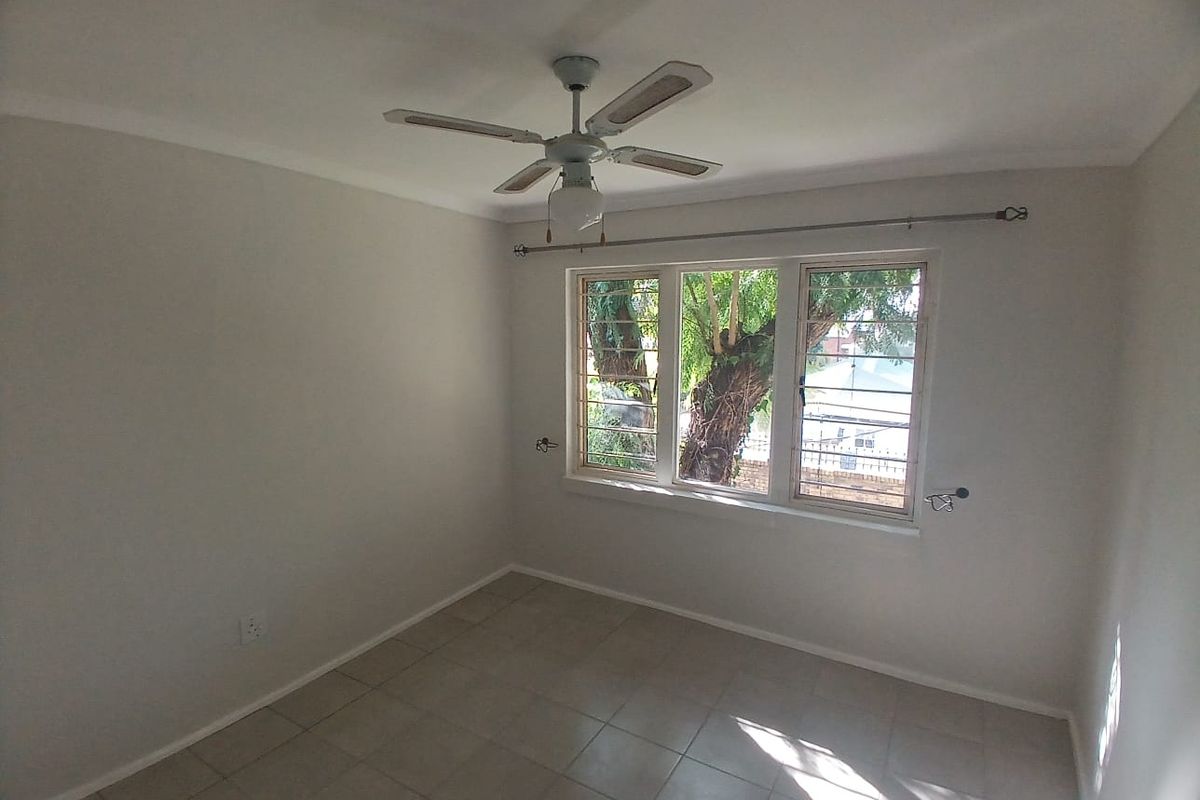 A bright, empty bedroom featuring neutral tiled flooring, a ceiling fan, and large windows fitted with security bars.