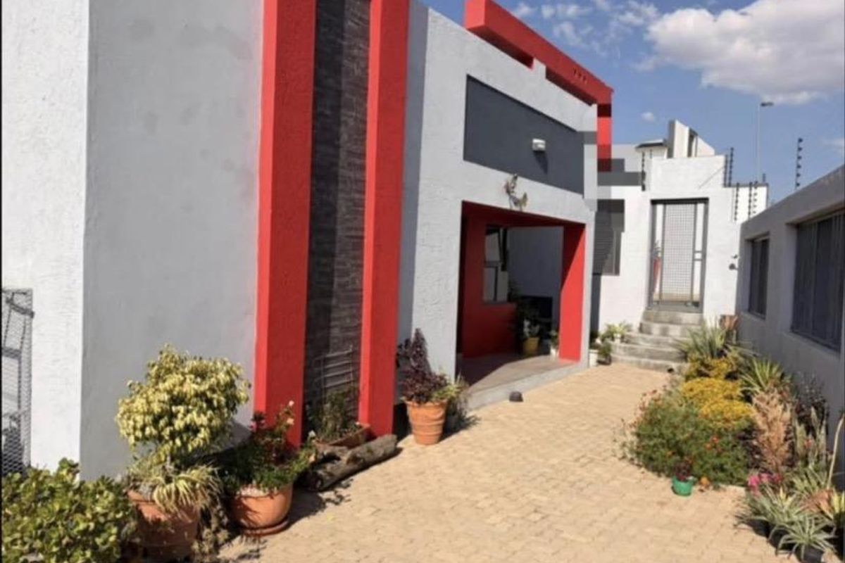 A modern rear exterior view featuring a paved courtyard, covered patio area, and distinctive red and grey architectural accents with security fencing.
